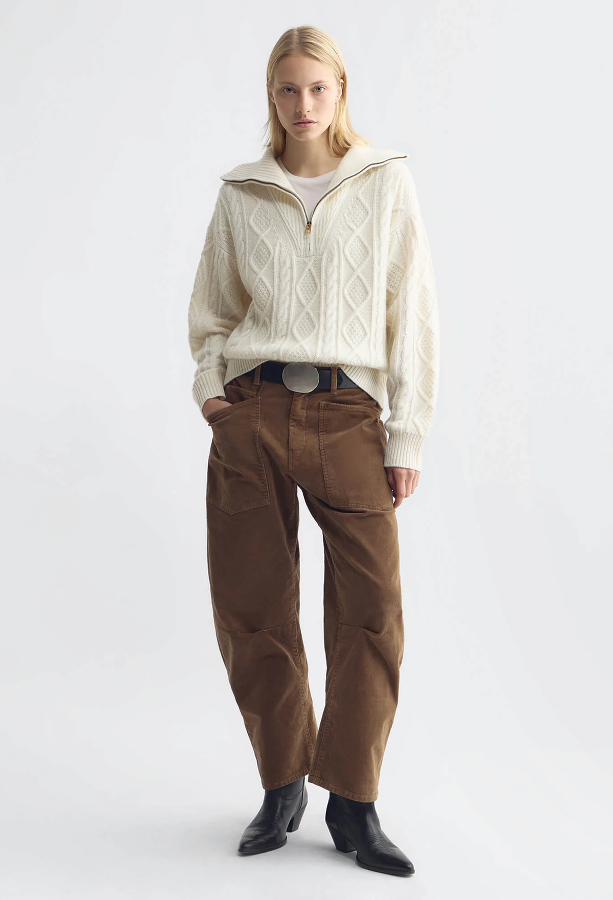 A model stands against a light gray studio background wearing a cream cable-knit half-zip sweater, brown corduroy pants, and black ankle boots.