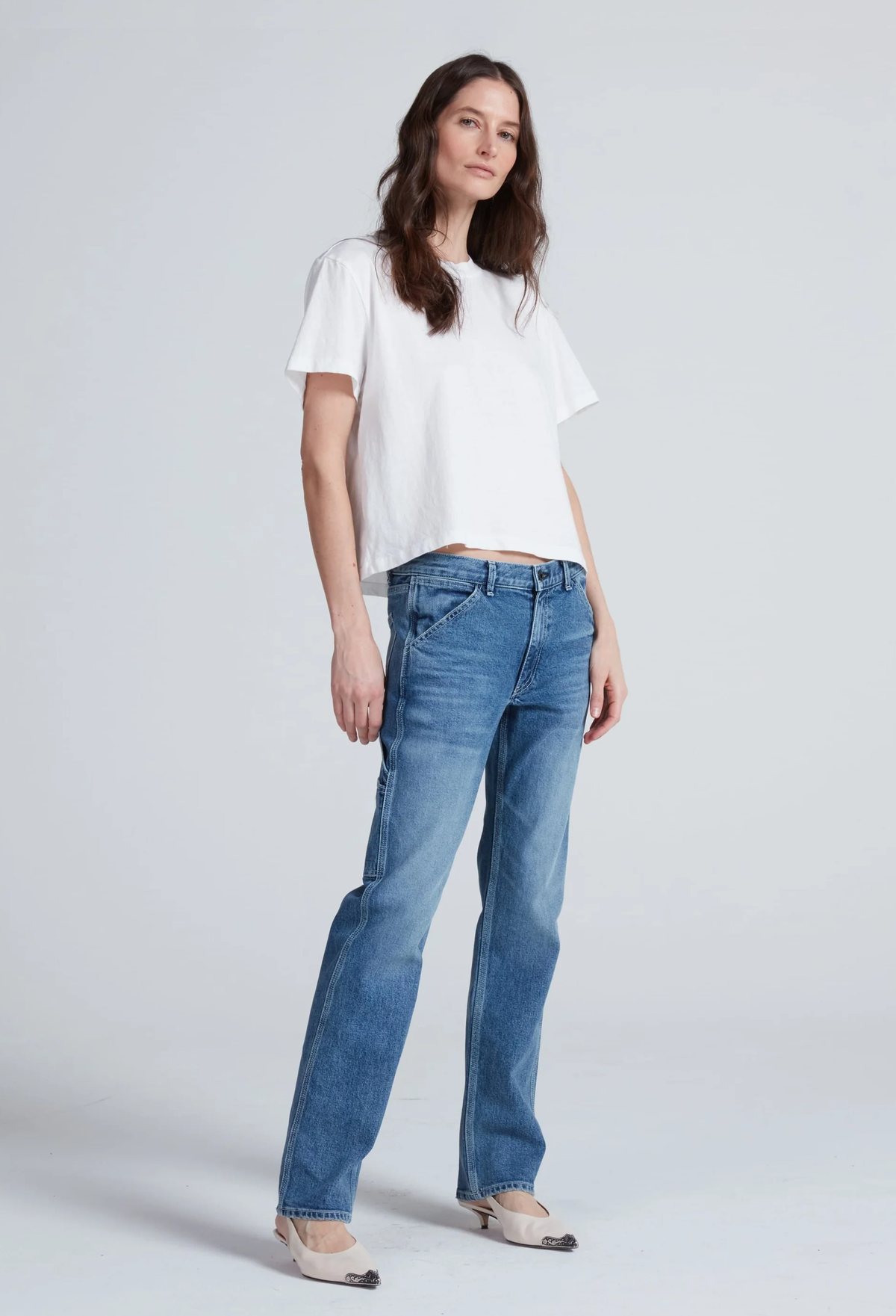A woman modeling a white short-sleeve crop top and blue straight-leg jeans against a light gray studio background.