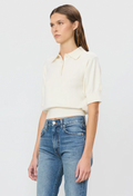 A model wearing a cream-colored short-sleeve polo shirt tucked into blue jeans, photographed against a clean, light gray studio background.