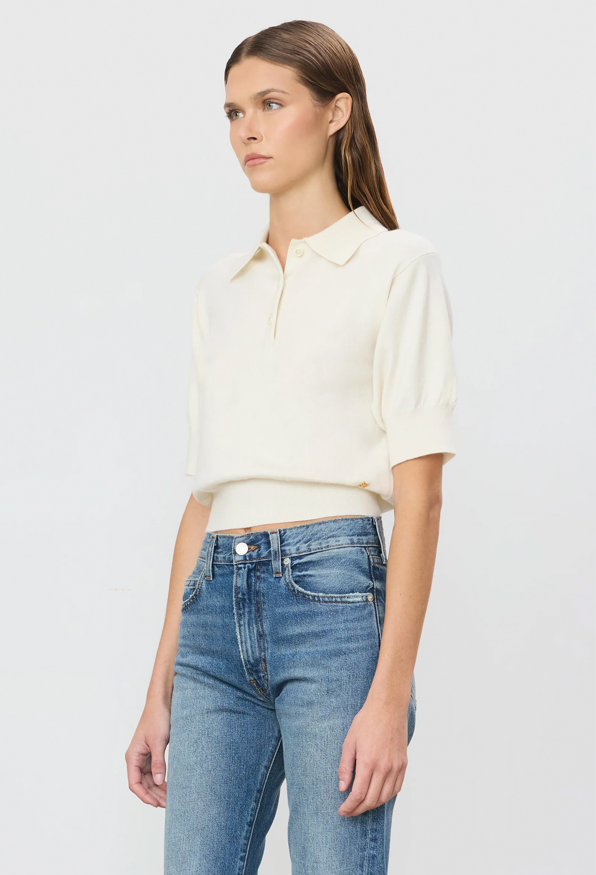 A model wearing a cream-colored short-sleeve polo shirt tucked into blue jeans, photographed against a clean, light gray studio background.