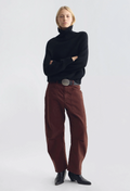 A model stands against a light gray studio background wearing a black turtleneck sweater, brown wide-leg pants with a silver belt, and black ankle boots.