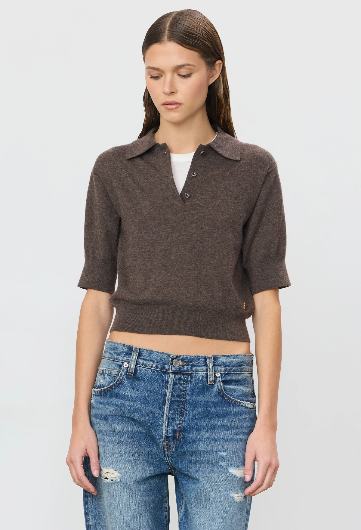 A woman modeling a short-sleeve brown knit polo crop top over a white undershirt, paired with blue distressed jeans, against a light gray studio background.