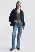 A model stands against a light gray studio background wearing a black fringed jacket, a gray lace-trimmed top, blue jeans, and black pointed shoes.