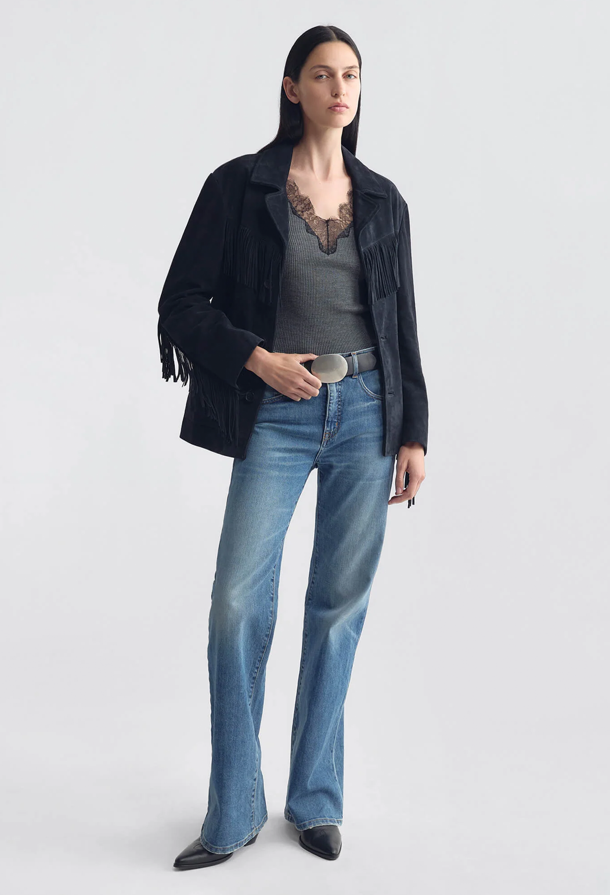 A model stands against a light gray studio background wearing a black fringed jacket, a gray lace-trimmed top, blue jeans, and black pointed shoes.