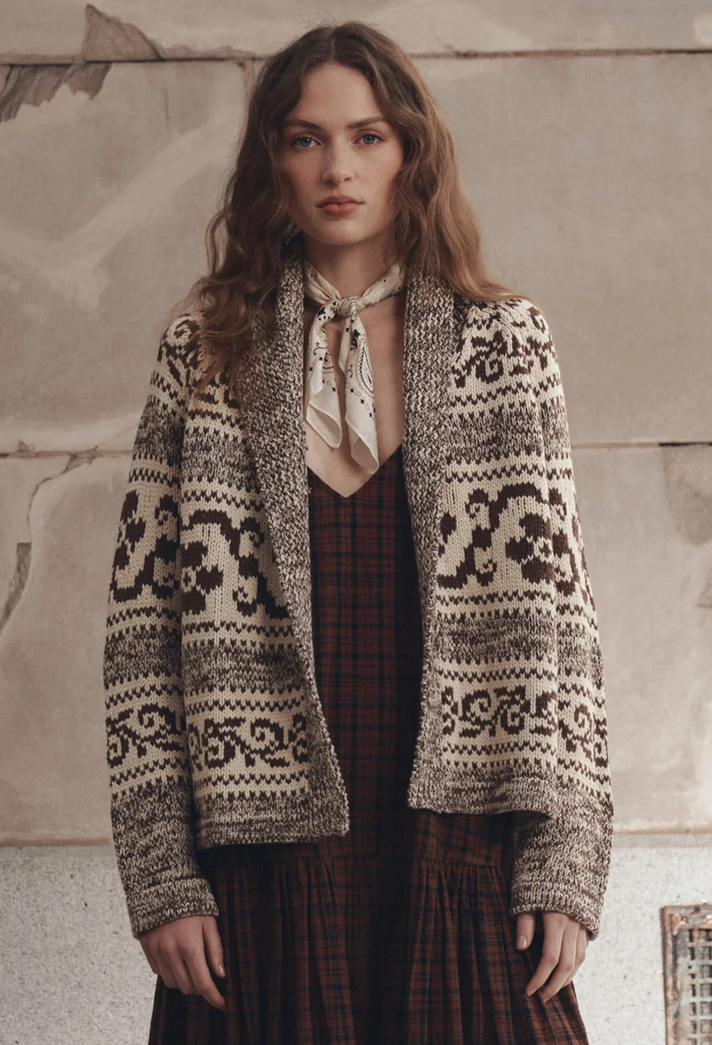 A woman wearing a cream and dark brown patterned knit cardigan over a red plaid dress, with a cream neck scarf, standing against a textured stone wall.