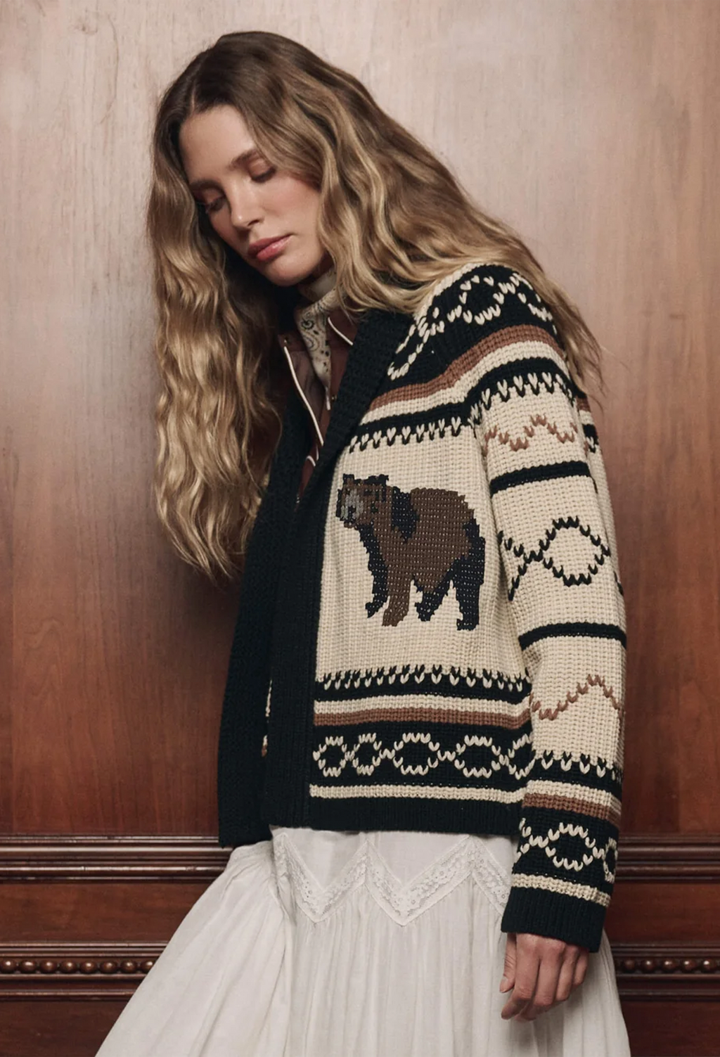 A woman with long wavy blonde hair wears a cream knit cardigan featuring a brown bear motif and dark geometric patterns, posed against a warm wood-paneled wall.