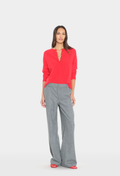 A woman modeling a bright red long-sleeve knit top with a split V-neckline and gray wide-leg trousers, standing against a light gray studio background.