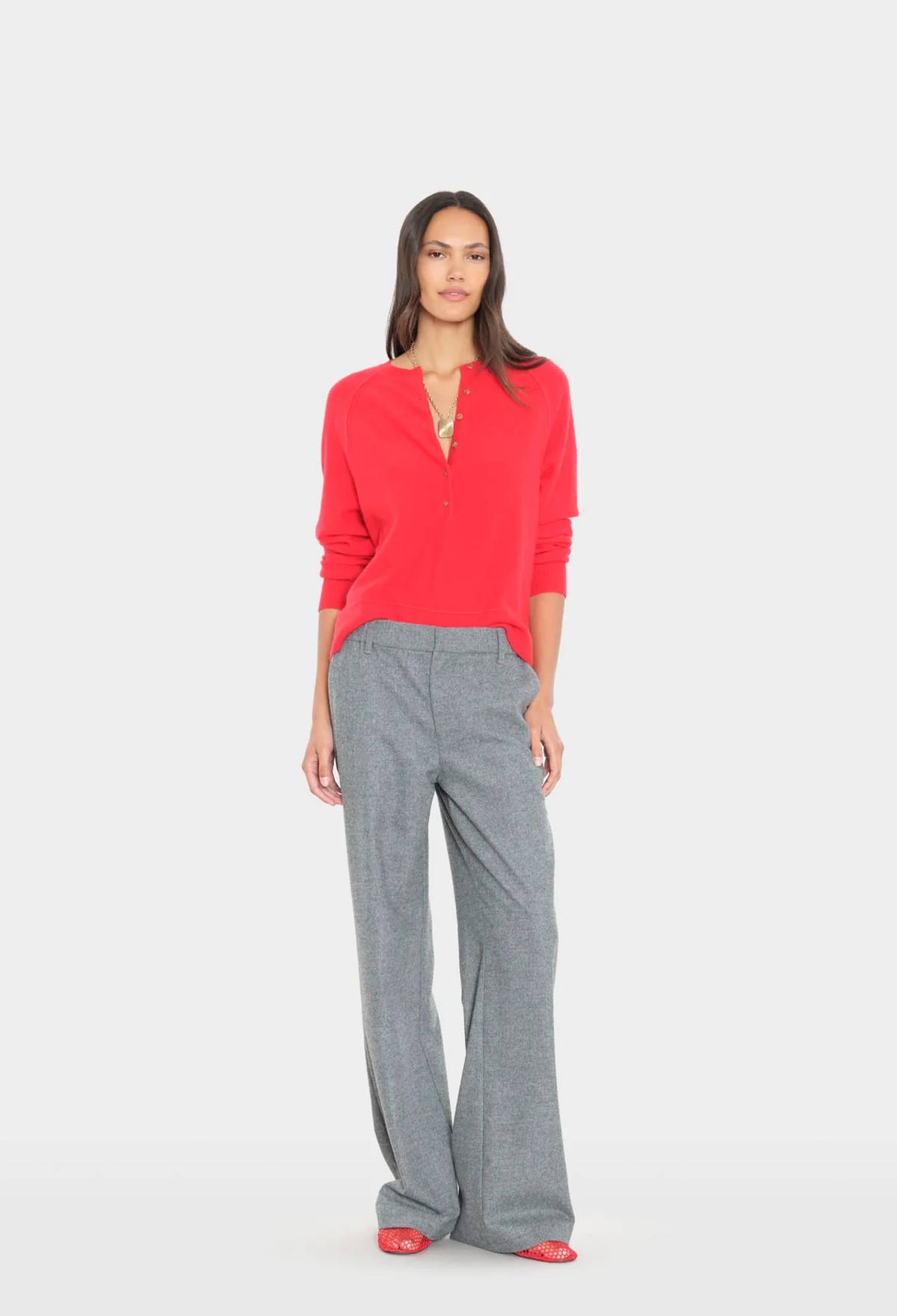 A woman modeling a bright red long-sleeve knit top with a split V-neckline and gray wide-leg trousers, standing against a light gray studio background.