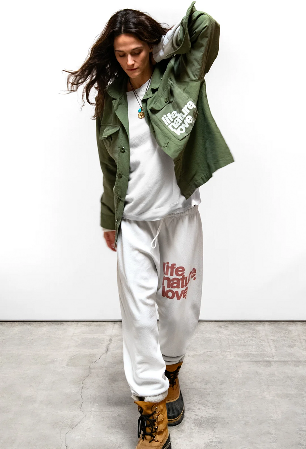 A woman modeling an olive green military-style jacket with white graphic text, worn over a white sweatshirt and white sweatpants with red text, paired with brown lace-up boots, against a white wall and concrete floor.