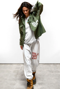 A woman modeling an olive green military-style jacket with white graphic text, worn over a white sweatshirt and white sweatpants with red text, paired with brown lace-up boots, against a white wall and concrete floor.
