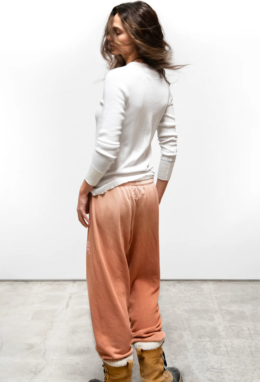 A woman stands in profile wearing a white long-sleeve knit top and loose peach-colored jogger pants with a white fuzzy cuff, paired with tan boots, against a plain white wall and concrete floor.