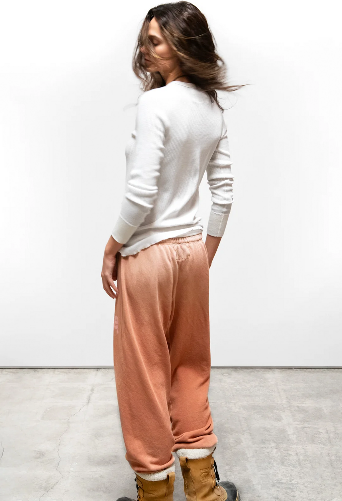 A woman stands in profile wearing a white long-sleeve knit top and loose peach-colored jogger pants with a white fuzzy cuff, paired with tan boots, against a plain white wall and concrete floor.
