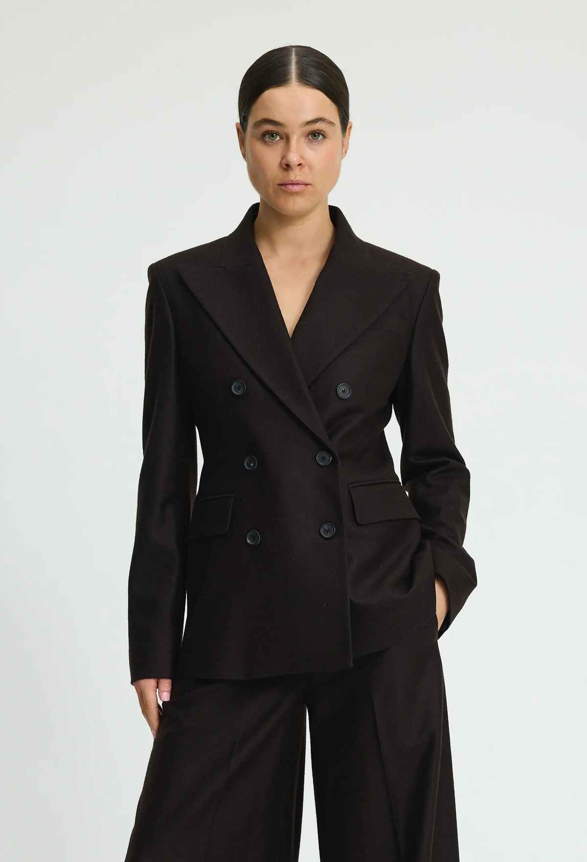 A woman modeling a dark brown double-breasted blazer with matching wide-leg trousers against a light gray studio background.