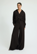 A woman modeling a black wide-leg jumpsuit with a matching belt and chest pocket details, photographed against a plain light gray studio background.