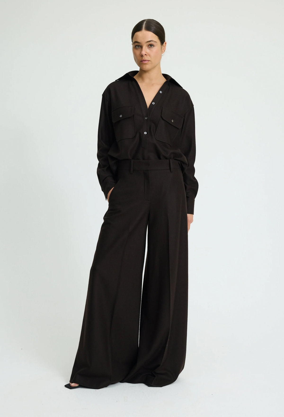 A woman modeling a black wide-leg jumpsuit with a matching belt and chest pocket details, photographed against a plain light gray studio background.