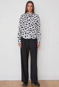 A model stands against a white wall wearing a black and white patterned long-sleeve blouse with black pleated wide-leg trousers and black shoes.