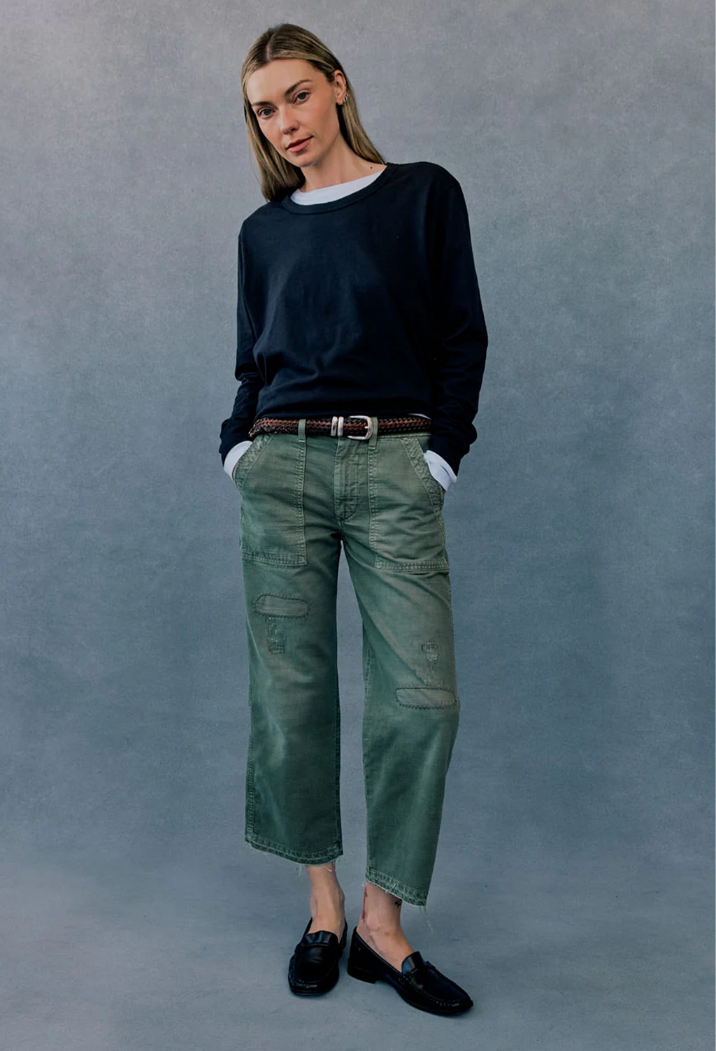 A woman modeling a pair of green cropped cargo-style jeans with a brown belt, paired with a dark navy long-sleeve top and black loafers, standing against a textured gray studio backdrop.