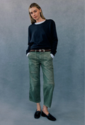 A woman modeling a pair of green cropped cargo-style jeans with a brown belt, paired with a dark navy long-sleeve top and black loafers, standing against a textured gray studio backdrop.
