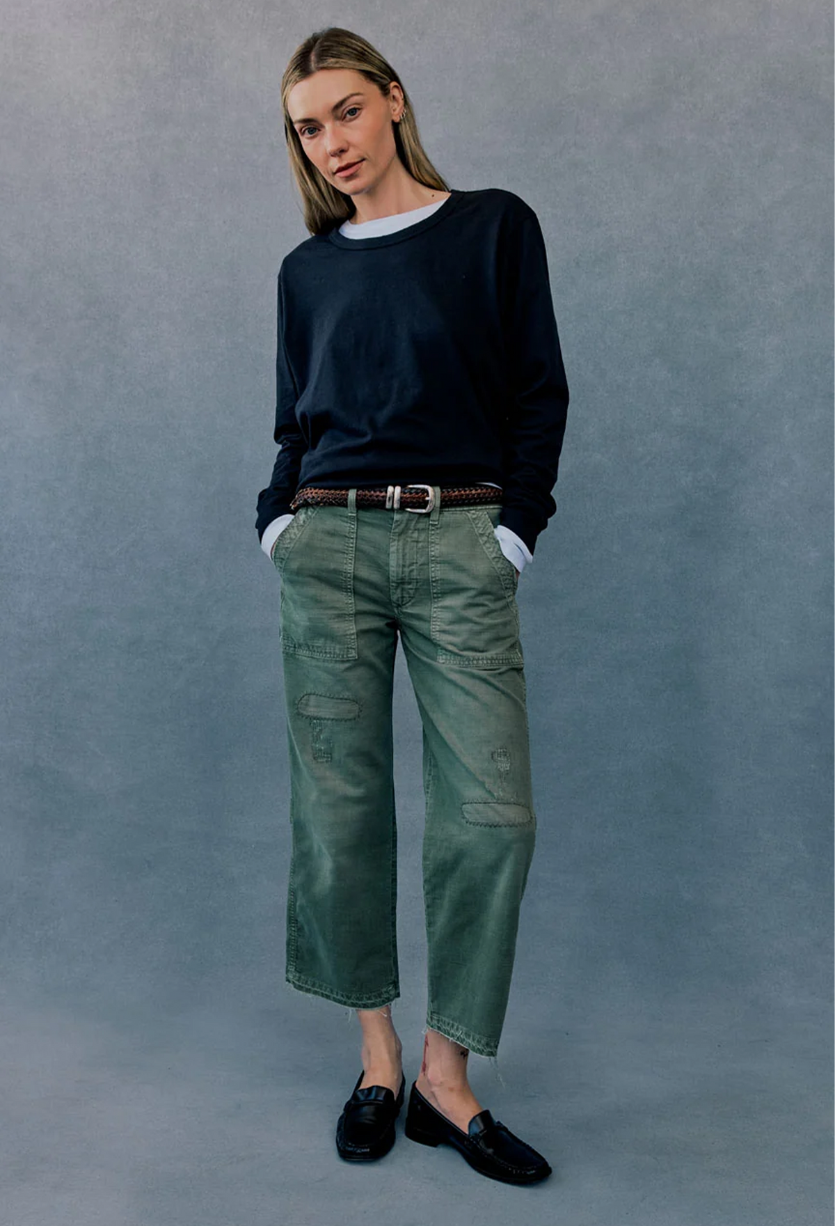 A woman modeling a pair of green cropped cargo-style jeans with a brown belt, paired with a dark navy long-sleeve top and black loafers, standing against a textured gray studio backdrop.