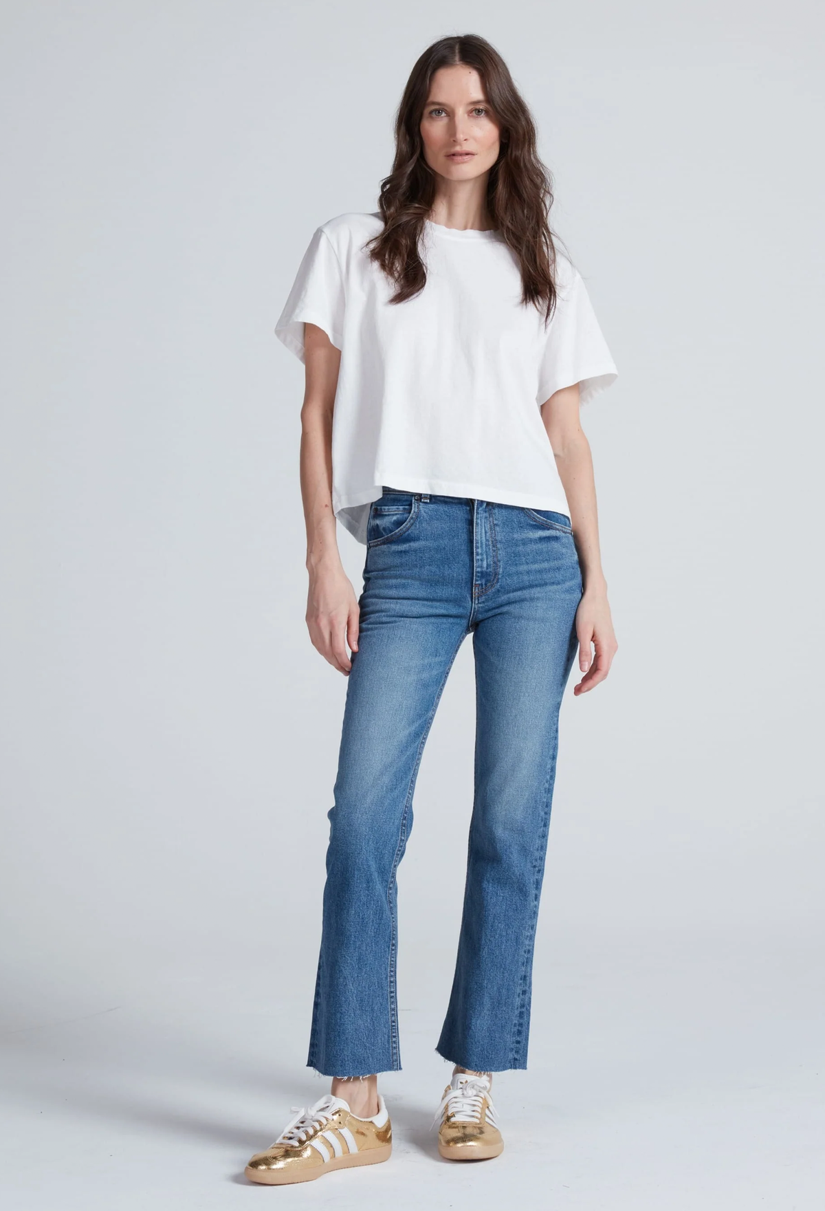 A woman modeling a white short-sleeve crop top and blue flare jeans with gold sneakers, standing against a light gray studio background.