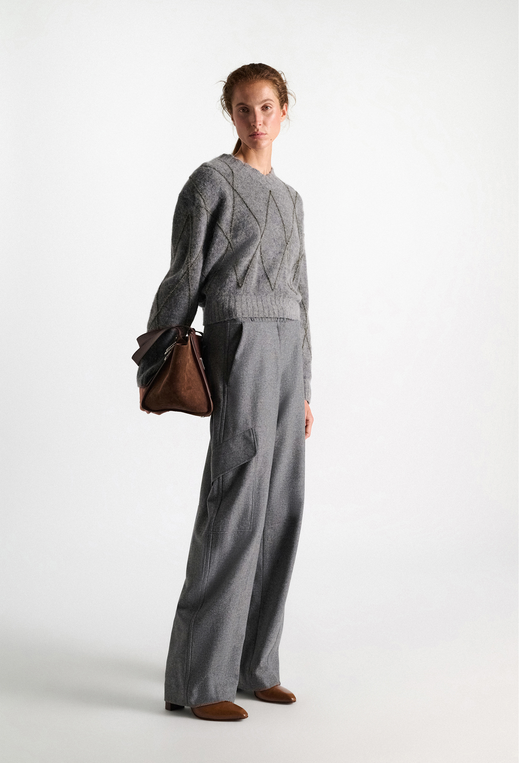 A model stands in a studio wearing a gray knit sweater and matching wide-leg trousers, holding a brown leather handbag.