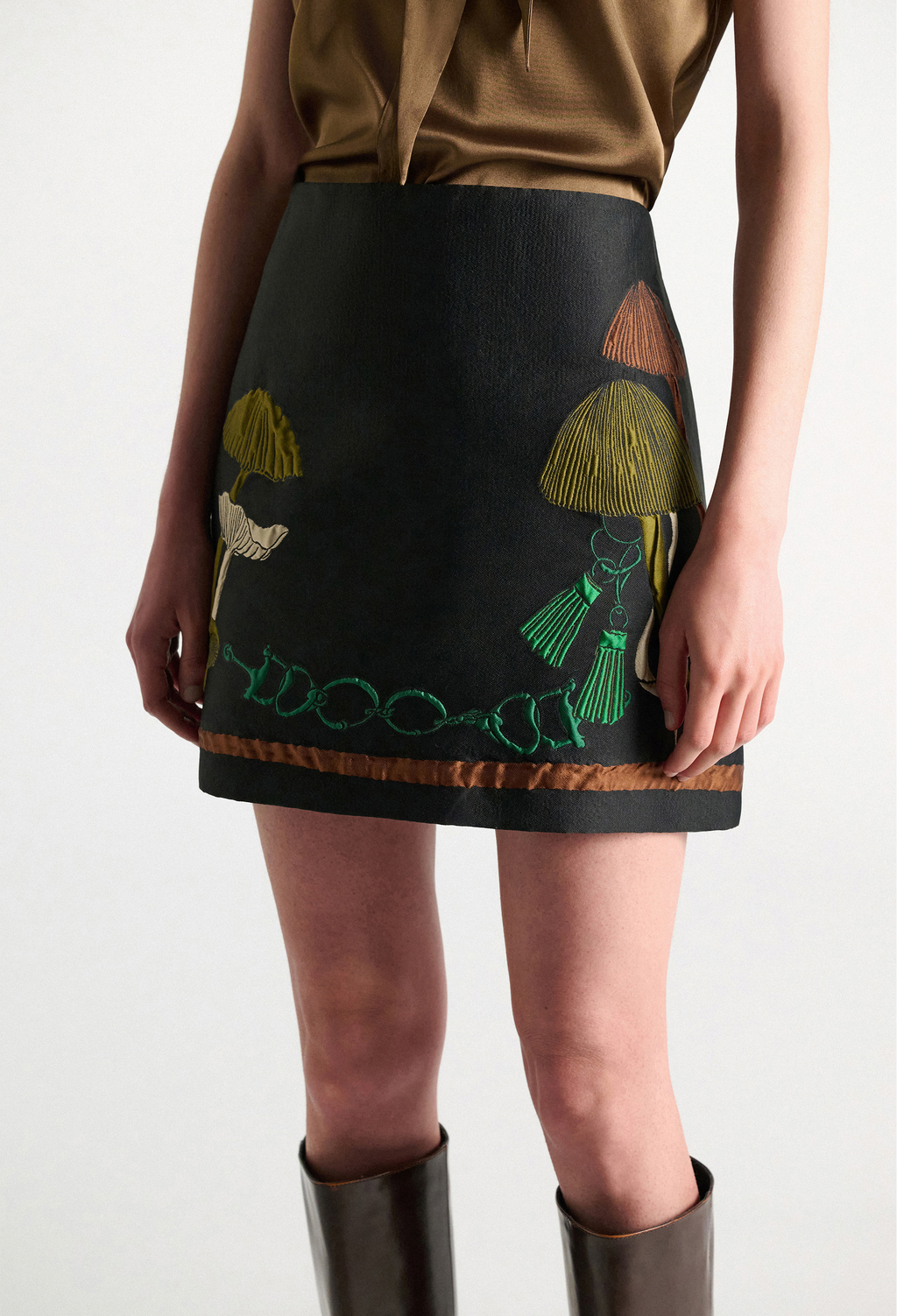 A model wearing a black mini skirt with embroidered mushroom motifs in green, gold, and brown, styled with a brown top and dark boots against a plain white background.