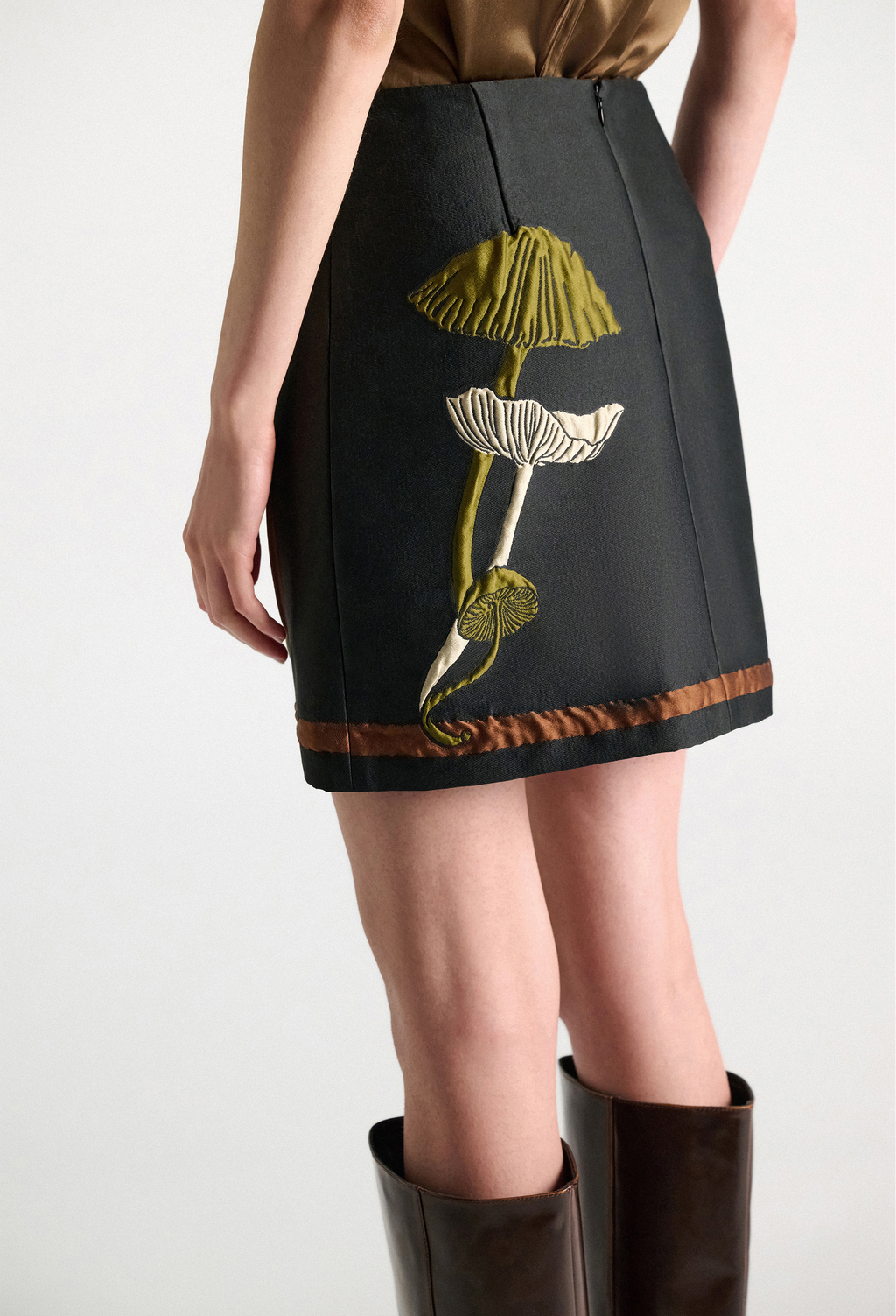 A model wearing a black A-line skirt with a large embroidered mushroom design on the front, paired with dark brown leather knee-high boots against a light gray studio background.