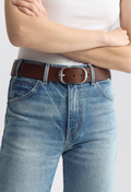 A close-up of a person wearing a brown leather belt with a silver-tone oval buckle, paired with blue jeans and a white top against a light gray studio background.