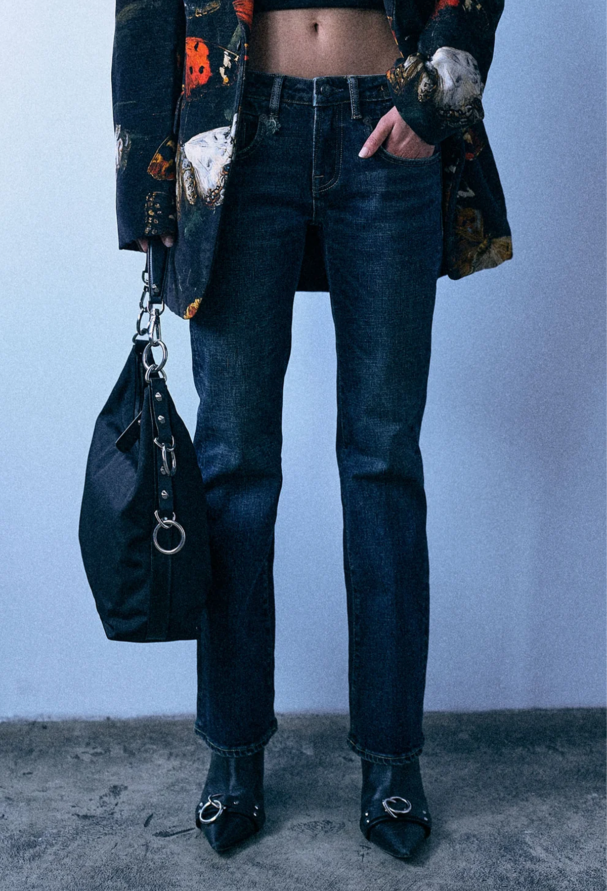 A person wearing dark blue straight-leg jeans, a dark floral-patterned jacket, and black pointed-toe ankle boots with silver rings, holding a black shoulder bag against a pale blue wall.