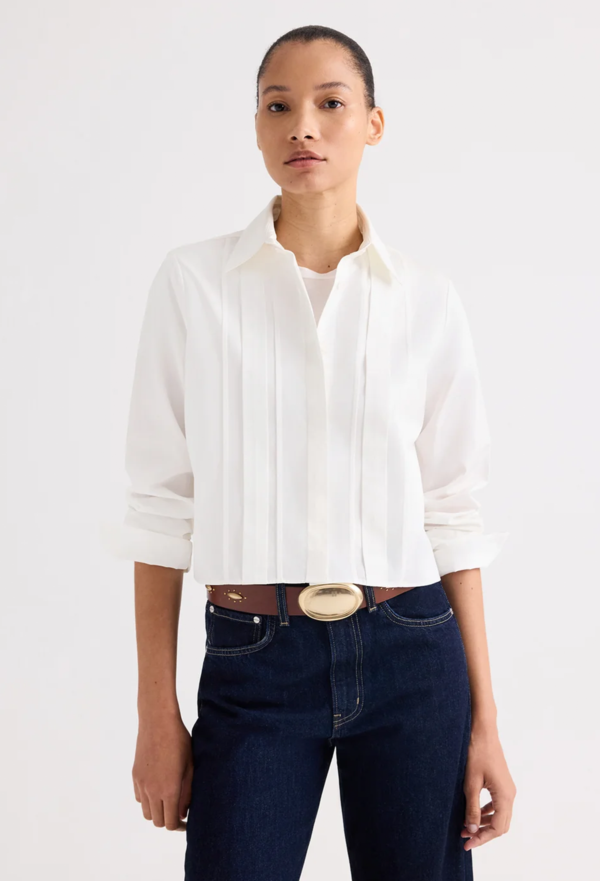 A model wearing a white long-sleeve pleated blouse tucked into dark blue jeans with a brown belt, photographed against a clean white studio background.