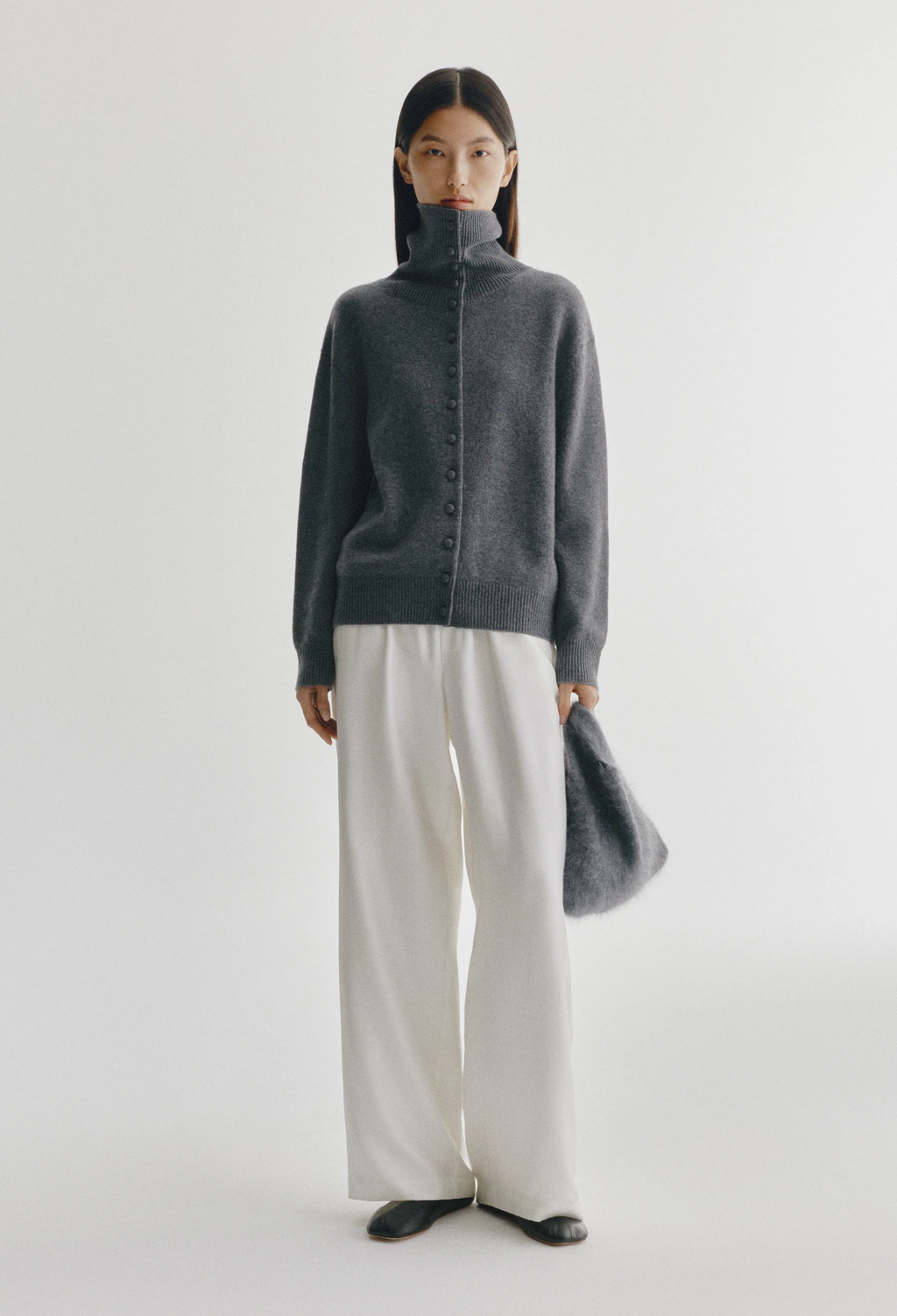 A model stands against a light gray studio background wearing a dark charcoal gray turtleneck button-front sweater, off-white wide-leg trousers, black shoes, and holding a matching dark gray bag.