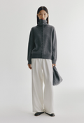 A model stands against a light gray studio background wearing a dark charcoal gray turtleneck button-front sweater, off-white wide-leg trousers, black shoes, and holding a matching dark gray bag.