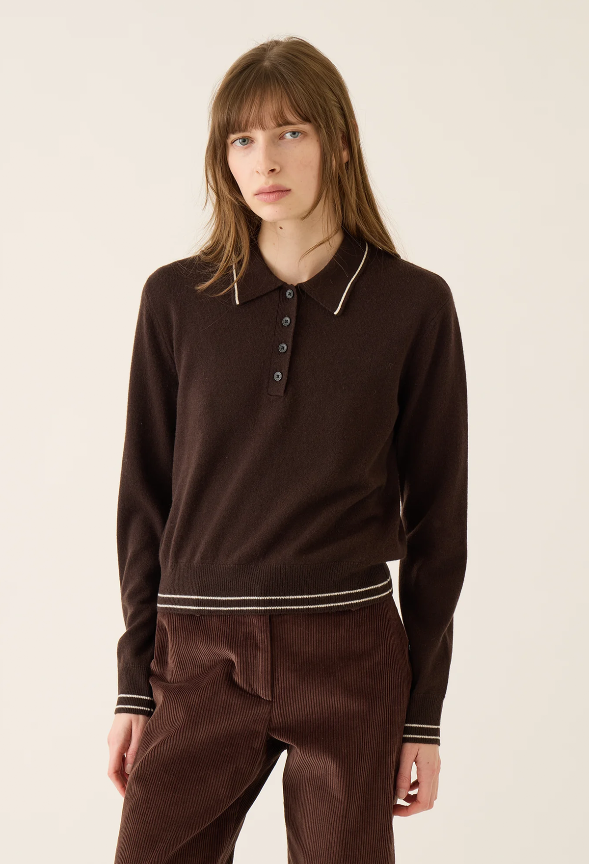 A model wearing a dark brown long-sleeve knit polo shirt with contrasting white piping on the collar and hem, paired with matching brown corduroy trousers, against a plain off-white studio background.