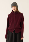 A model wearing a dark burgundy zip-up knit sweater with a mock neck, paired with a gray beanie and a black skirt, photographed against a plain off-white background.