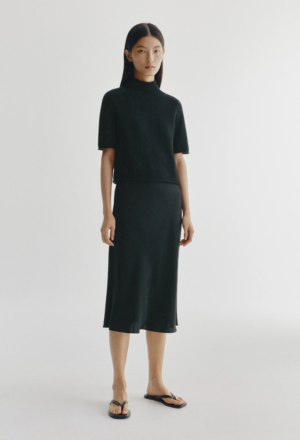 A model stands against a light gray studio background wearing a matching black knit two-piece set consisting of a short-sleeve mock-neck top and a midi skirt, paired with black sandals.