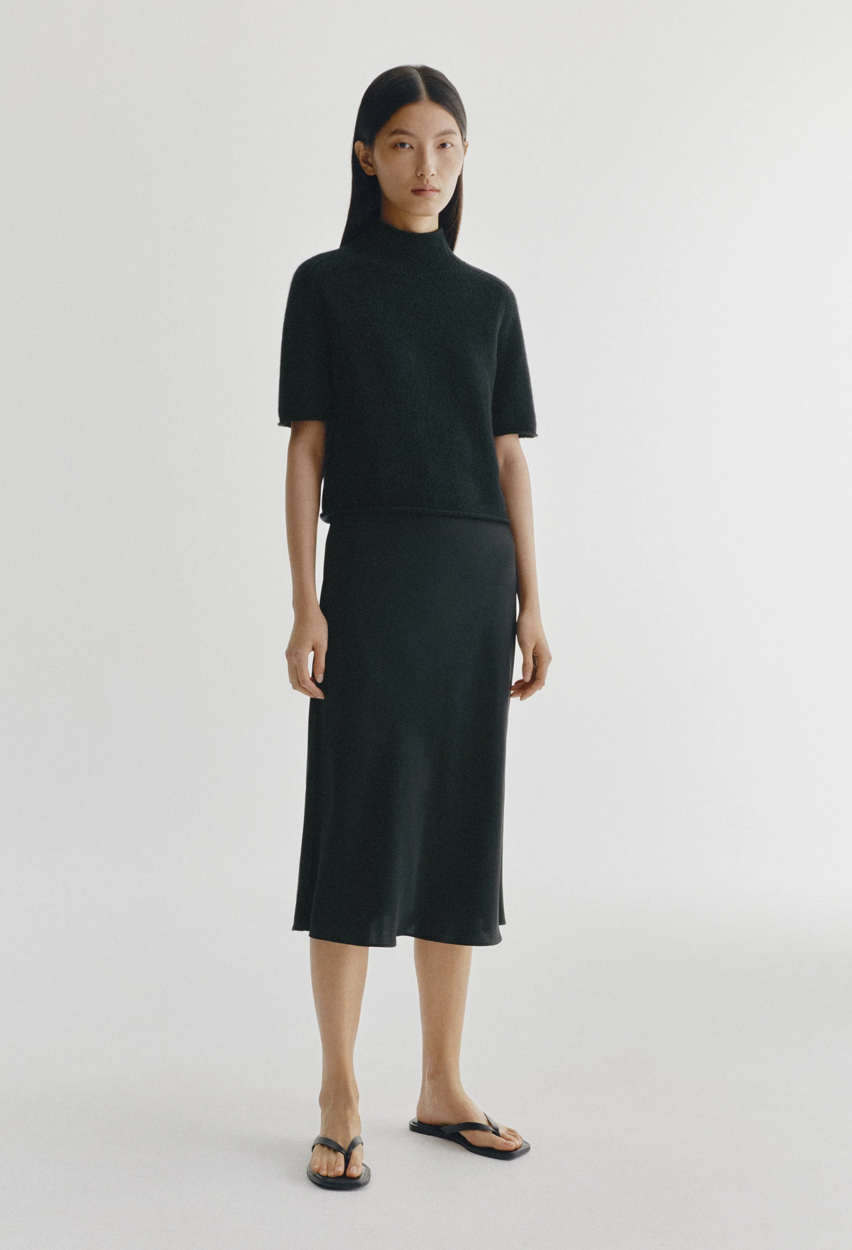 A model stands against a light gray studio background wearing a matching black knit two-piece set consisting of a short-sleeve mock-neck top and a midi skirt, paired with black sandals.