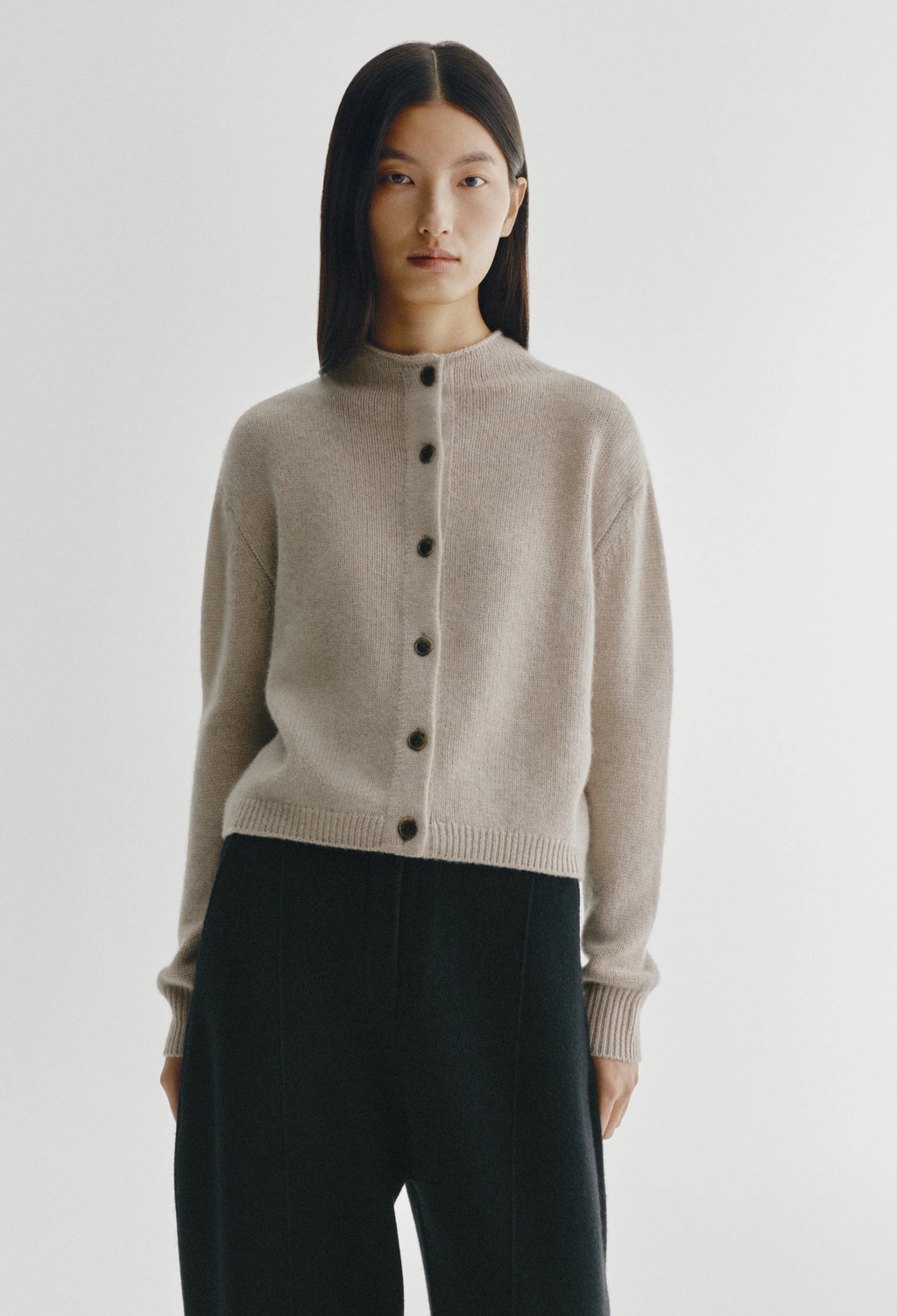 A model wearing a light beige knit cardigan with a button-up front and dark buttons, paired with dark charcoal pants, against a plain light gray studio background.