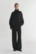 A model stands against a light gray studio background wearing a dark brown oversized turtleneck sweater and matching wide-leg trousers.