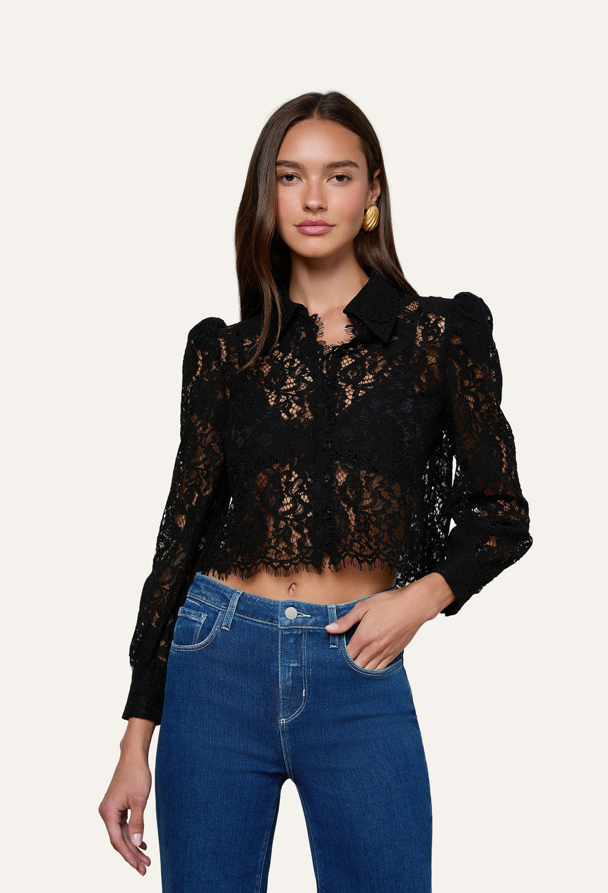 A woman modeling a black lace long-sleeve cropped blouse with puffed shoulders, paired with blue jeans, against a plain off-white studio background.