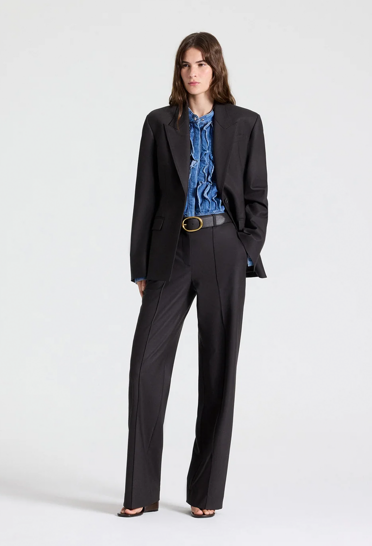 A model stands against a light gray studio background wearing a black tailored blazer and matching wide-leg trousers over a blue ruffled blouse, with a black belt and black sandals.