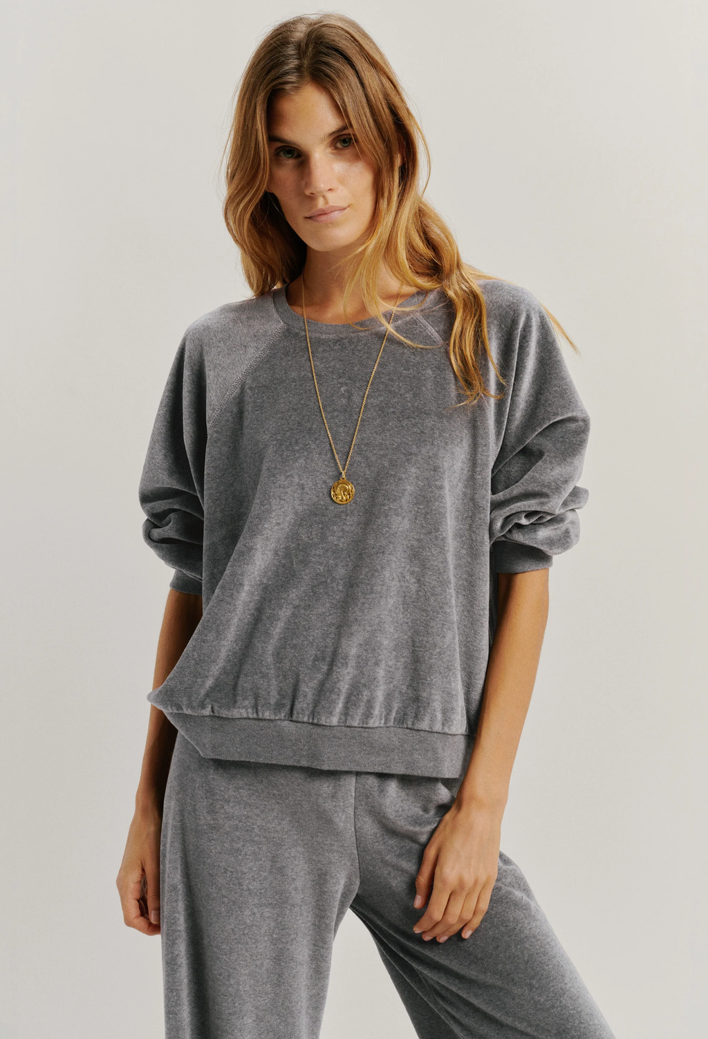 A woman modeling a matching gray velour lounge set, featuring a long-sleeve sweatshirt with gathered cuffs and matching sweatpants, accessorized with a gold necklace against a light gray studio background.