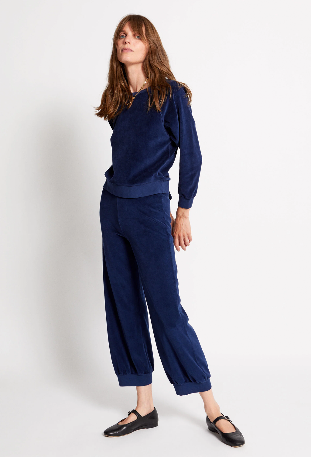 A woman modeling a matching navy blue velour lounge set consisting of a long-sleeve top and wide-leg cropped pants, paired with black Mary Jane shoes against a plain white studio background.