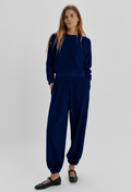 A woman modeling a matching navy blue loungewear set consisting of a long-sleeve crewneck sweatshirt and relaxed-fit jogger pants with elastic cuffs, paired with black Mary Jane flats, against a light gray studio background.