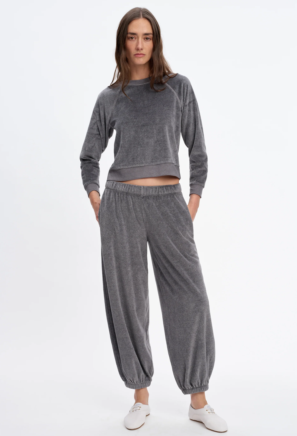 A woman modeling a matching gray velour lounge set consisting of a cropped long-sleeve top and relaxed jogger pants, paired with white sneakers against a plain white studio background.