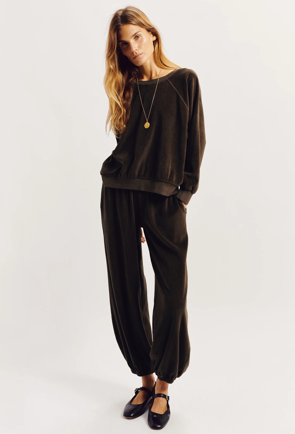 A woman modeling a dark brown velvet lounge set consisting of a long-sleeve sweatshirt and matching jogger pants, paired with black Mary Jane shoes and a gold pendant necklace, against a plain light gray background.