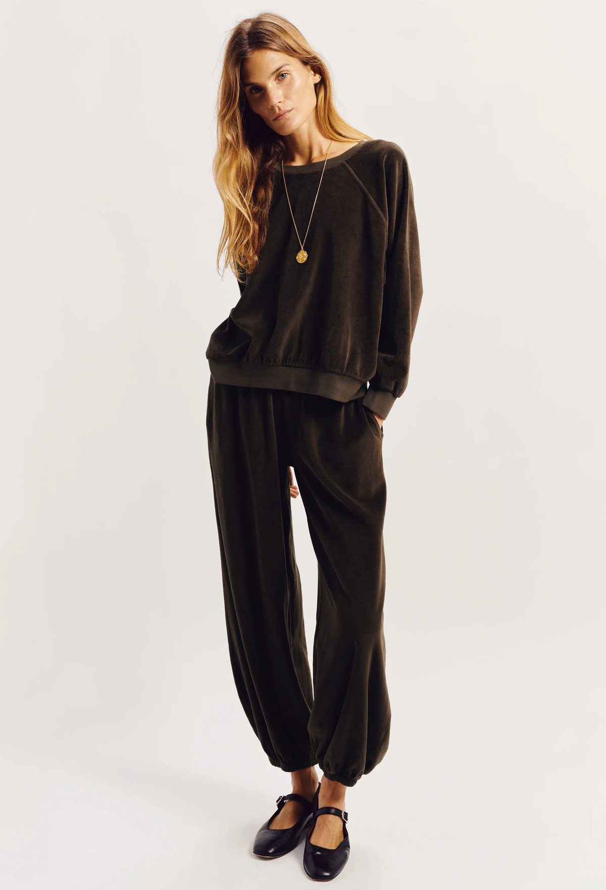 A woman modeling a dark brown velvet lounge set consisting of a long-sleeve sweatshirt and matching jogger pants, paired with black Mary Jane shoes and a gold pendant necklace, against a plain light gray background.