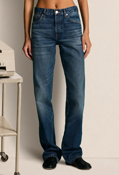 A model wearing a pair of medium-wash blue straight-leg jeans, standing in front of a light beige background with a vintage typewriter on a table to the side.