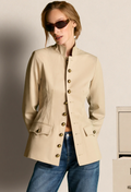 A woman wearing a beige cropped jacket with gold-tone buttons and blue jeans, standing against a light neutral wall.