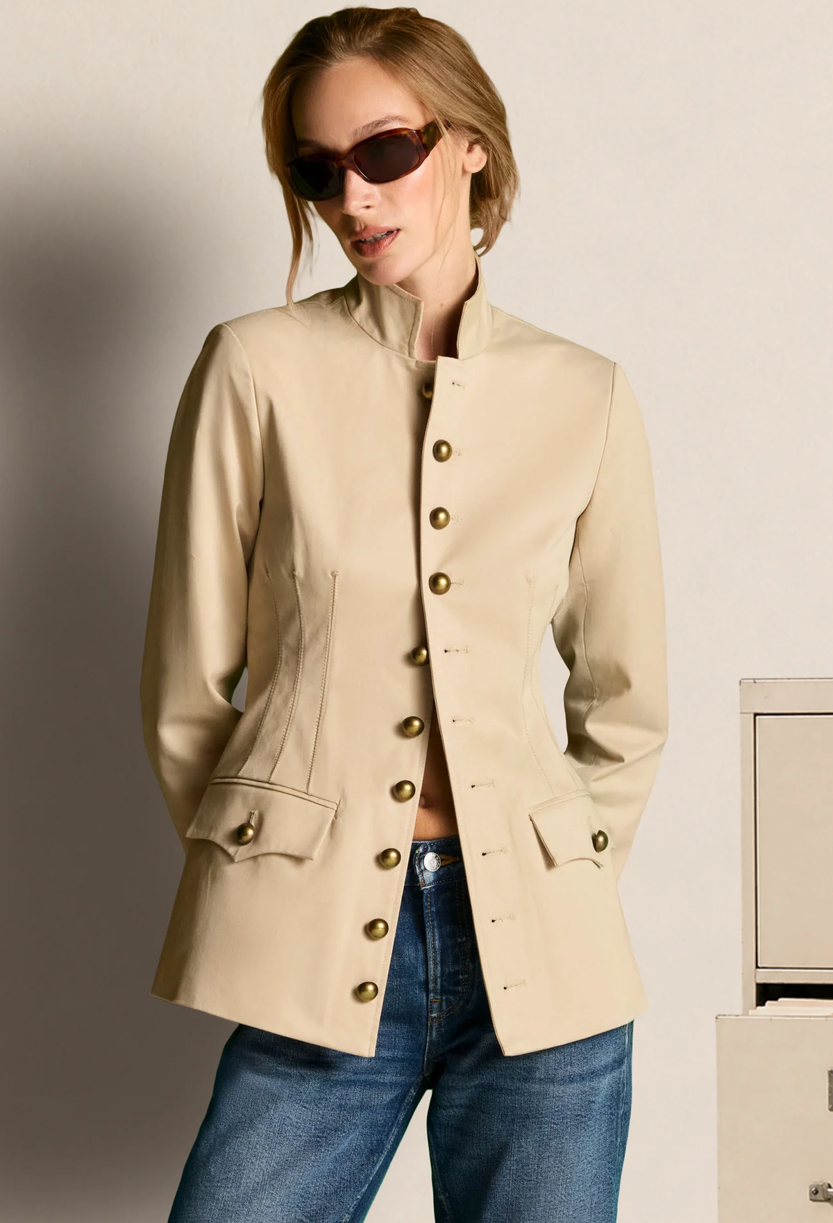 A woman wearing a beige cropped jacket with gold-tone buttons and blue jeans, standing against a light neutral wall.