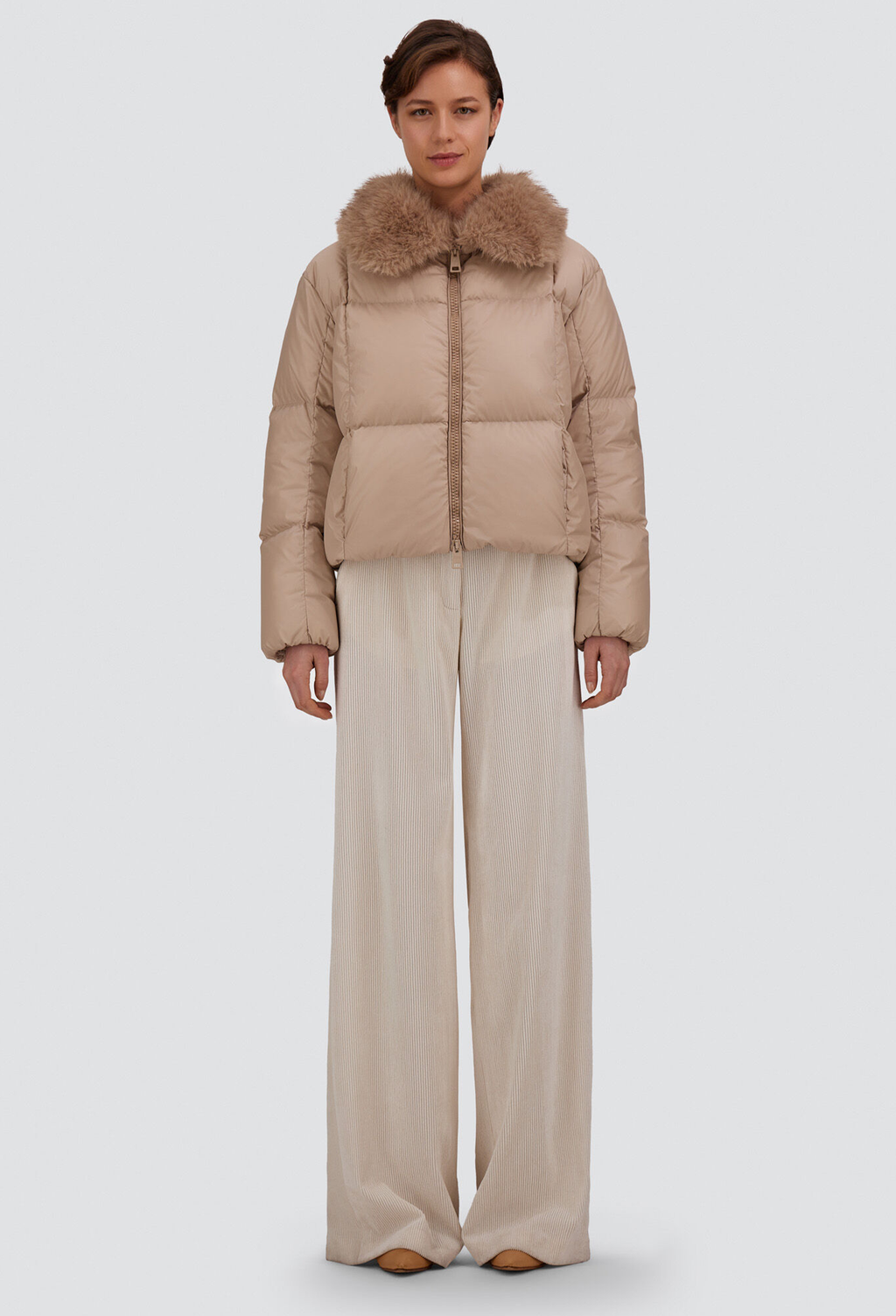 A model stands against a light gray studio background wearing a cropped beige puffer jacket with a large brown faux-fur collar and wide-leg beige trousers.